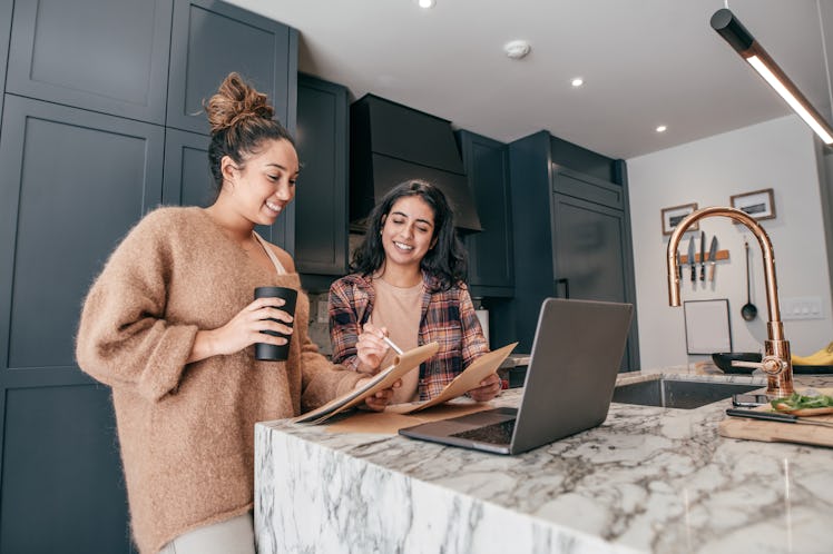 Two young women stand in a luxe kitchen and take notes while watching a video course on their laptop...