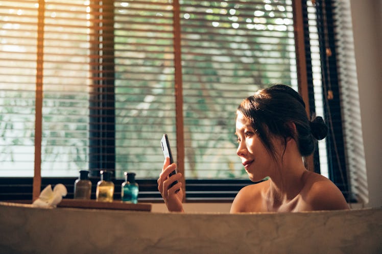 A young Asian woman sits in a bubble bath at golden hour and looks at her phone.