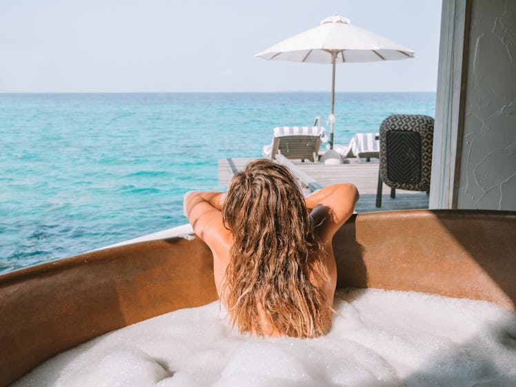 A young woman with blonde hair looks out at the ocean while in a bubble bath on vacation.