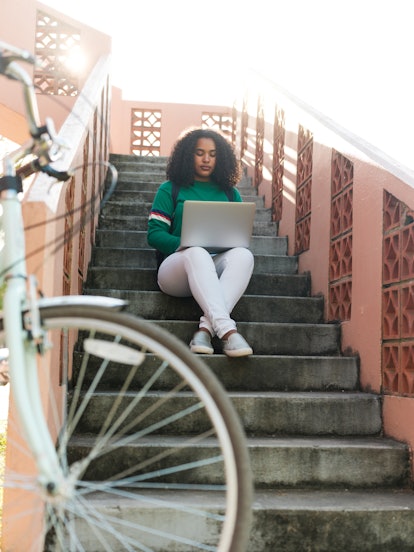 A young Black woman sits on an outdoor staircase and types on her laptop.