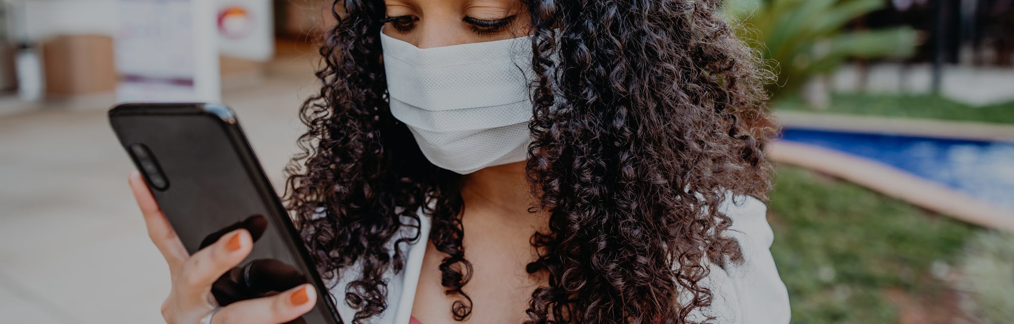 A woman with curly hair can be seen holding a smartphone while she wears a face mask in public.
