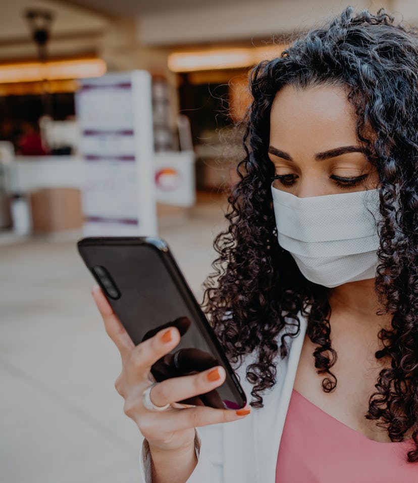 A woman with curly hair can be seen holding a smartphone while she wears a face mask in public.