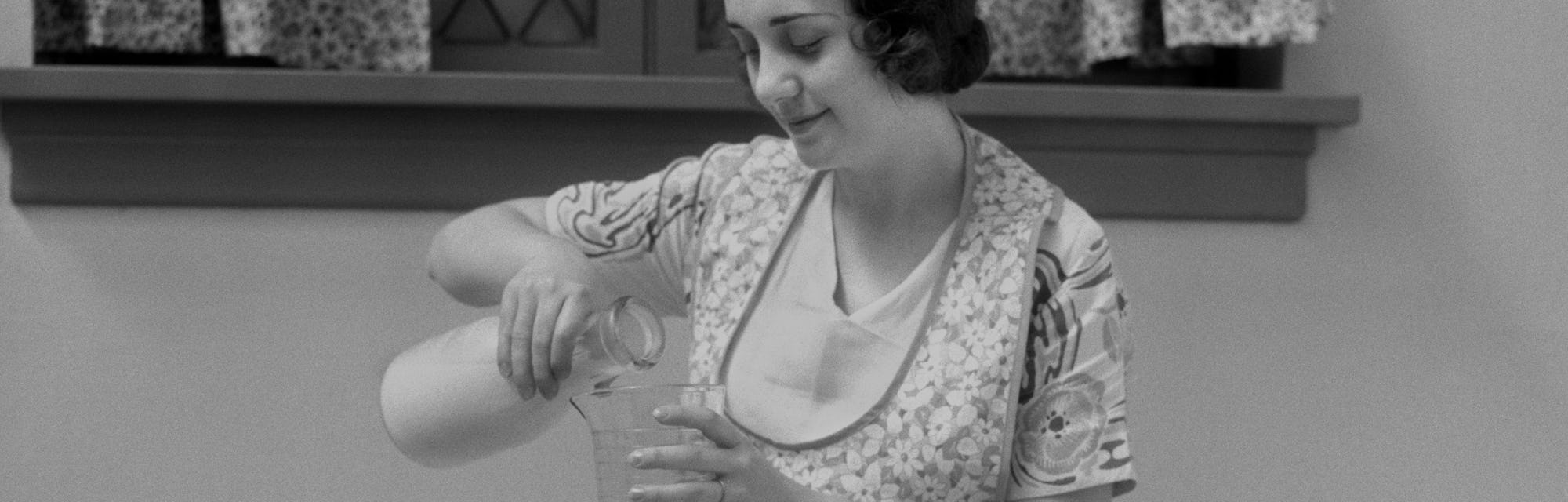 A 1920s image of a young woman preparing food at the table. She can be seen pouring milk from a jug ...