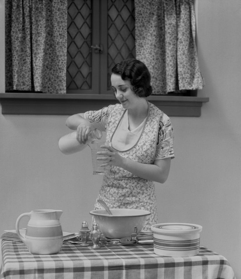 A 1920s image of a young woman preparing food at the table. She can be seen pouring milk from a jug ...