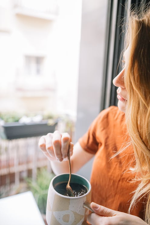 A blonde woman standing next to a window while holding a cup of tea during the quarantine