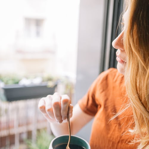 A blonde woman standing next to a window while holding a cup of tea during the quarantine