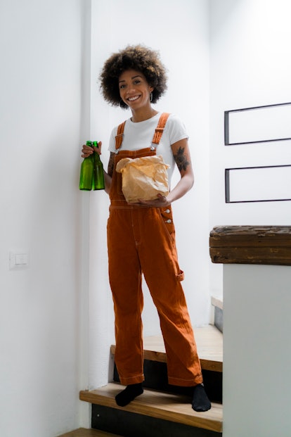 A young and stylish Black woman holds beer bottles and a bag of takeout food while posing on the sta...