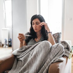 A woman in a blanket takes her temperature at home. Going to the doctor, dentist, and ob-gyn during ...