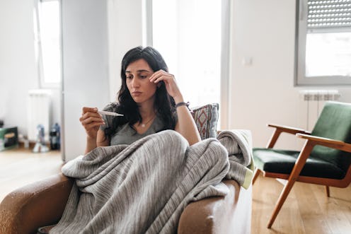 A woman in a blanket takes her temperature at home. Going to the doctor, dentist, and ob-gyn during ...