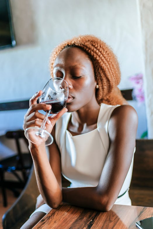 A young Black woman sits at a table and sips on a glass of red wine.