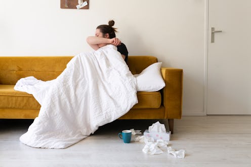 A woman coughs while sitting on a leather couch. What does a COVID-19 sore throat feel like? Doctors...