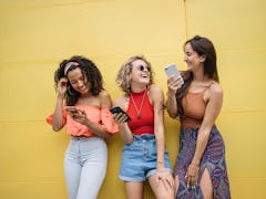 Three best friends laugh at something on their phones, while standing next to a yellow wall. 