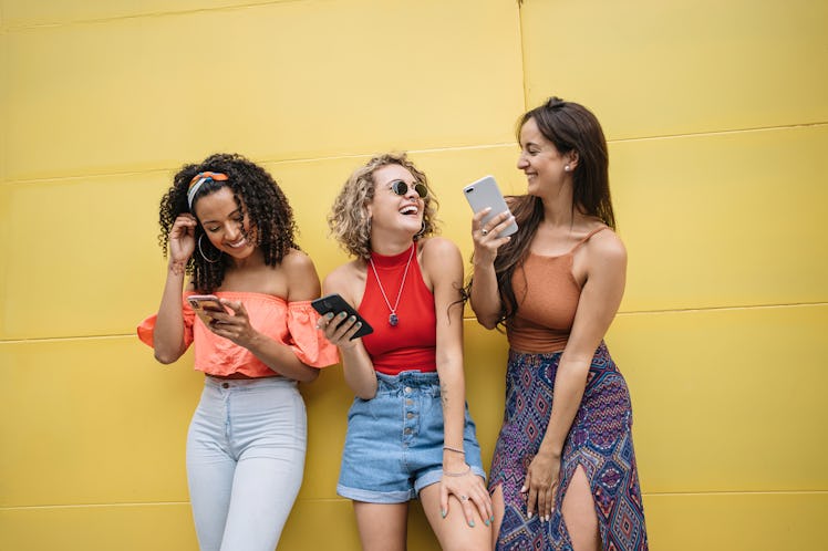 Three best friends laugh at something on their phones, while standing next to a yellow wall. 