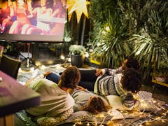 Two young couples lay on a palette with a blanket and lights on it, and watch a movie outdoors on a ...
