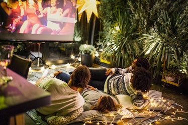 Two young couples lay on a palette with a blanket and lights on it, and watch a movie outdoors on a ...