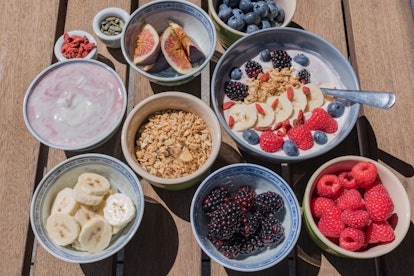 A smoothie bowl sits on a picnic table along with ingredients like fresh fruit, seeds, and granola t...