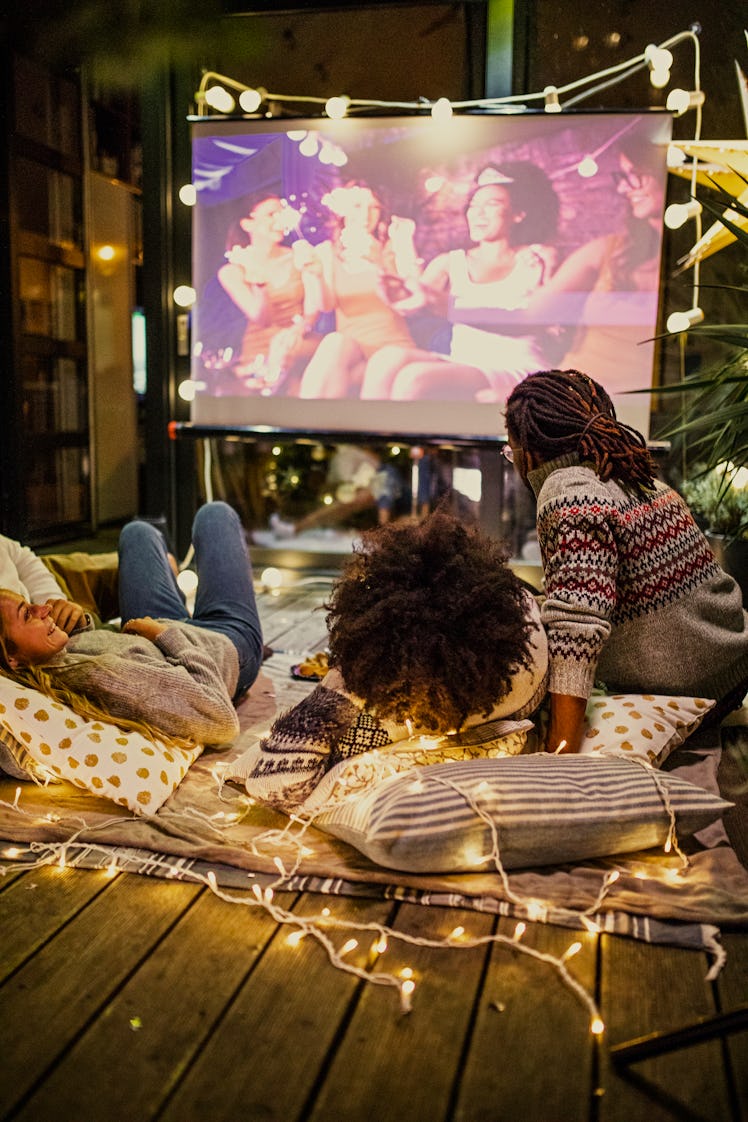 Two couples sit on a blanket that's lined with lights and watch a movie on a projected screen.