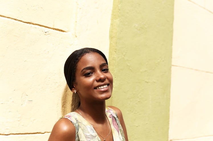 A young woman poses against a light yellow wall on a sunny day.