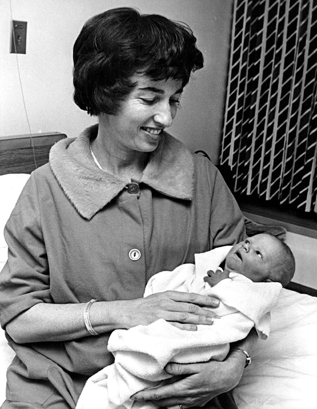 A mom dressed in her coat cradles her newborn in this vintage maternity ward photo.