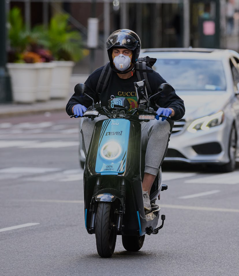 A rider with a helmet and face mask on can be seen riding a Revel scooter. Behind him is a car.
