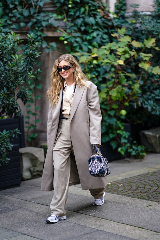 A woman in a beige shirt, grey coat, beige pants, white sneakers and white-blue bag