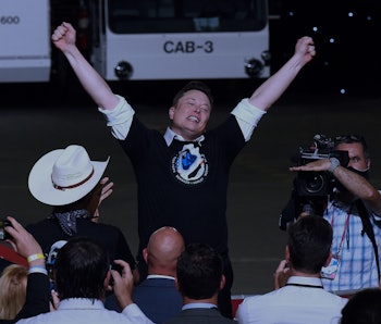 Elon Musk, wearing a SpaceX T-shirt, cheers with both arms up. 