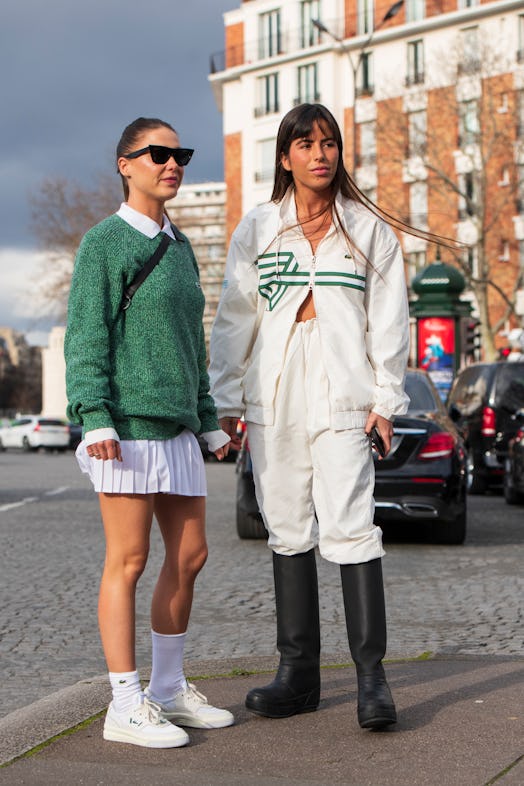 A woman in a white shirt, skirt and sneakers and green sweater and a woman in a white tracksuit and ...