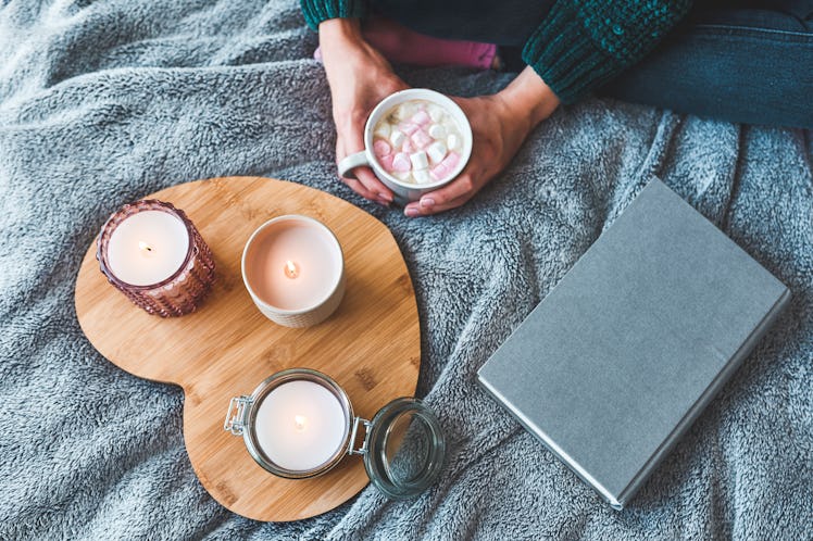 A young woman sits with a mug of hot cocoa, blanket, book, and three candles on heart-shaped wooden ...