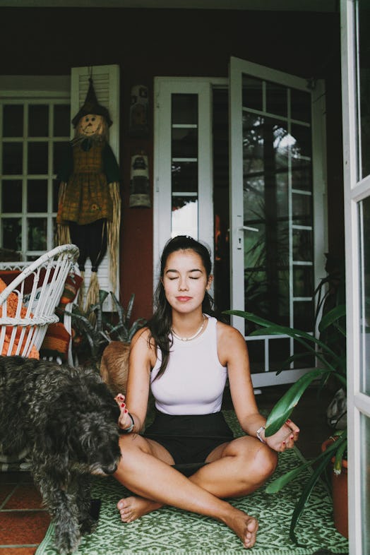 A young Asian woman sits on her porch and meditates on a rug while sitting next to her dog.