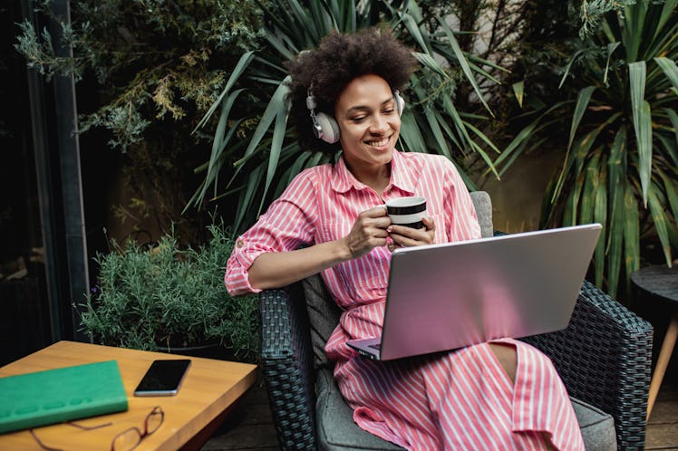 A young Black woman sits with her laptop on her back patio that's surrounded with plants and palm le...