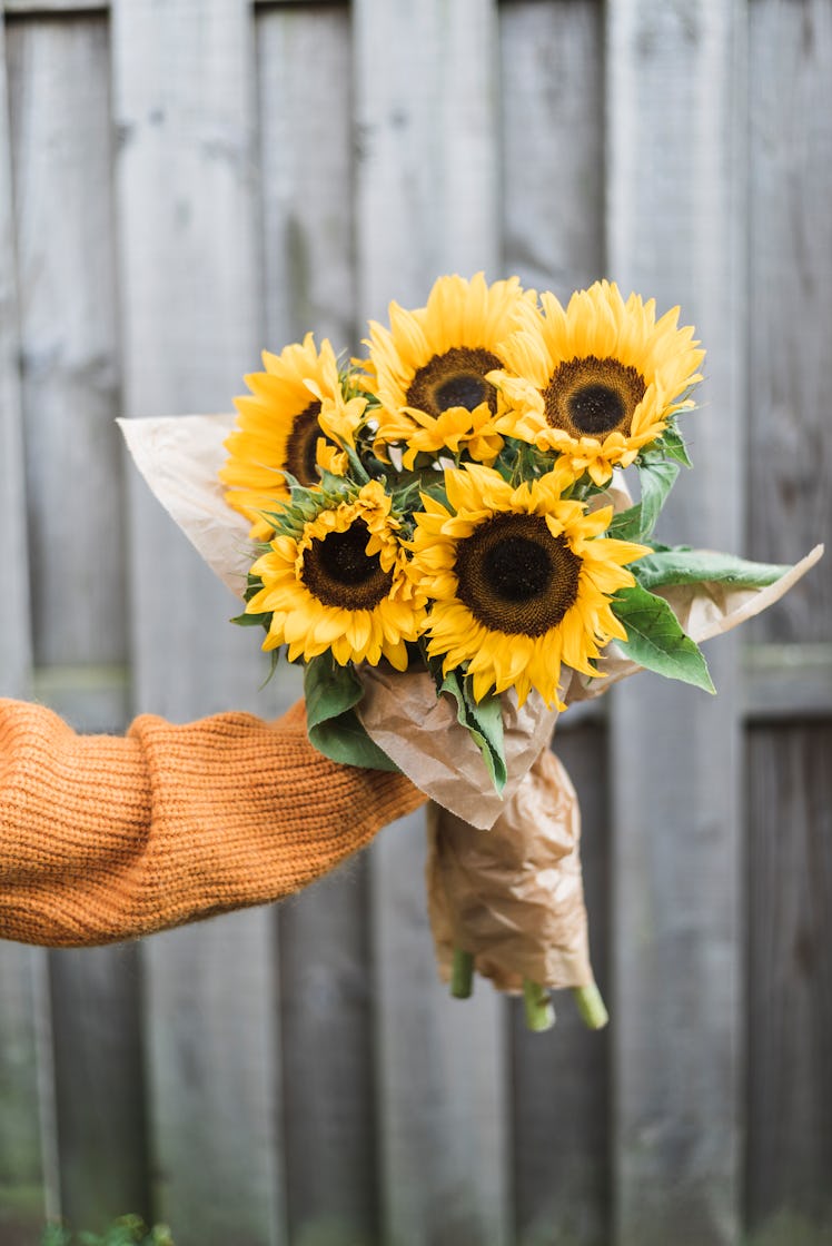 A young woman wearing an orange sweater holds up a bouquet of sunflowers in her backyard.