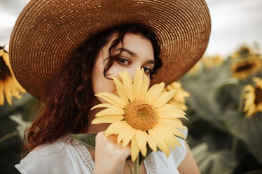 A young woman wearing a large sun hat holds a sunflower up to her mouth while posing for a picture.