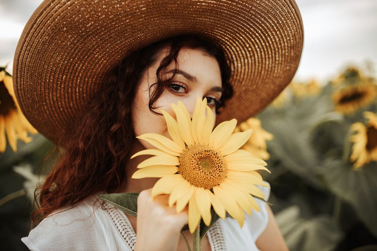 A young woman wearing a large sun hat holds a sunflower up to her mouth while posing for a picture.