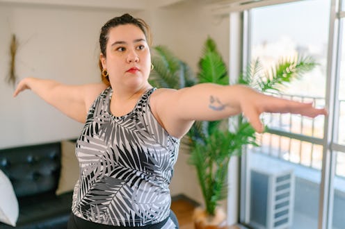 A person wearing a black and white yoga tank with a tattoo on her forearm poses in Warrior II. Findi...