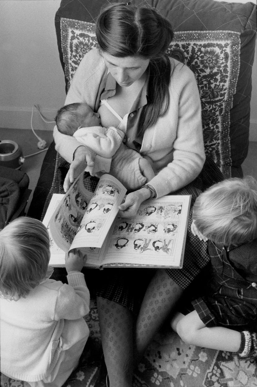 woman breastfeeding baby on lap while reading a book to two toddlers at her feet