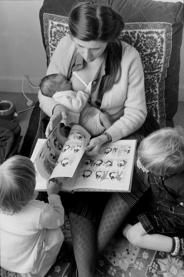 woman breastfeeding baby on lap while reading a book to two toddlers at her feet