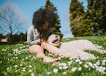A young Black woman sits in the grass in her backyard with her fluffy dog on a sunny day.