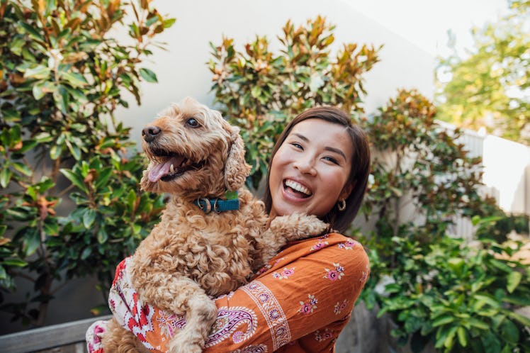 A young Asian woman holds her golden doodle and smiles brightly while standing amongst the plants in...