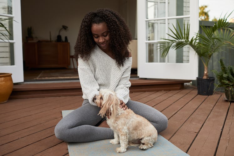 A young Black woman sits on a yoga mat on her deck and pets her pup.