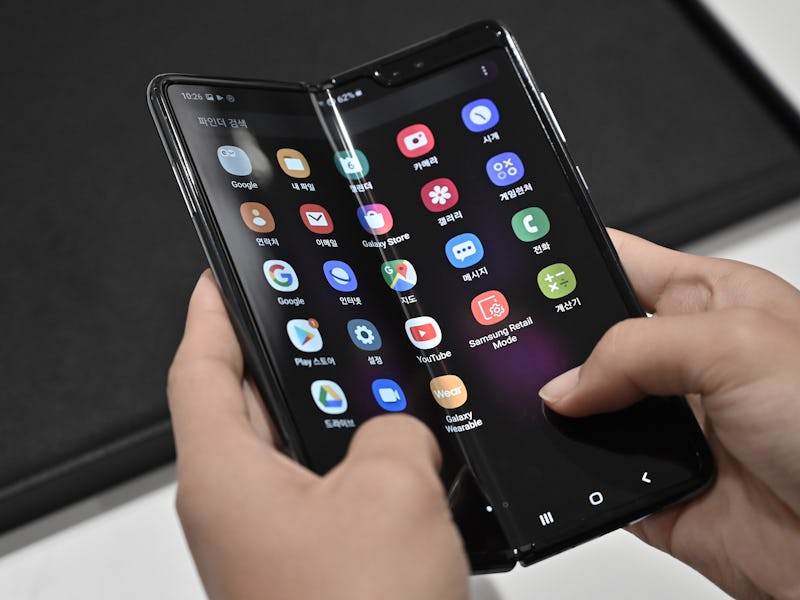 A foldable smartphone can be seen held by a pair of hands. The screen displays multiple app icons.
