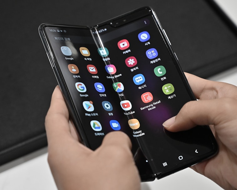 A foldable smartphone can be seen held by a pair of hands. The screen displays multiple app icons.