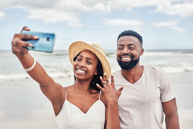 A young Black couple poses for a selfie after getting engaged on the beach.