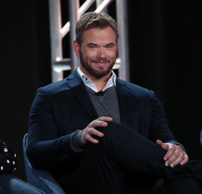 Kellan Lutz sits in a blue chair, smiling, with his legs crossed. He sports a full beard and is wear...