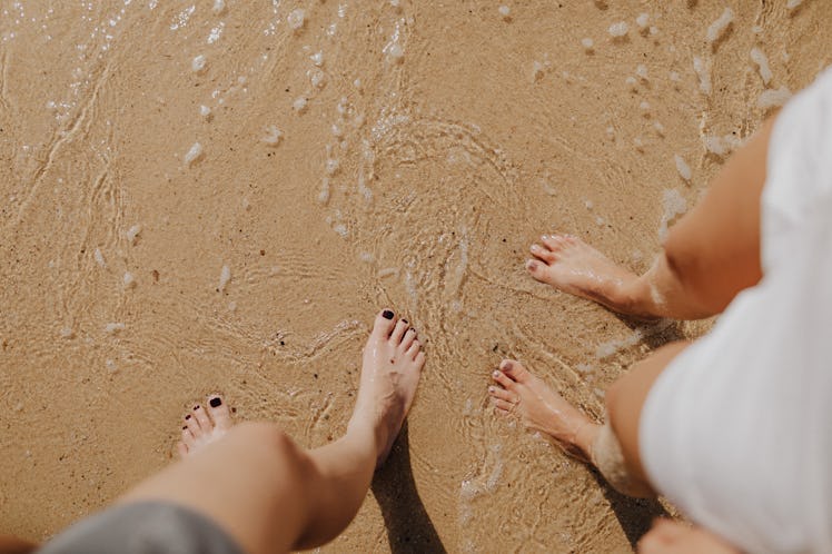 Two young women stand in the sand at the beach as the saltwater rushes back out to sea.