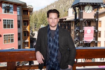 Peter Facinelli stands on a balcony in front of a pink apartment building. He wears a grey shirt and...