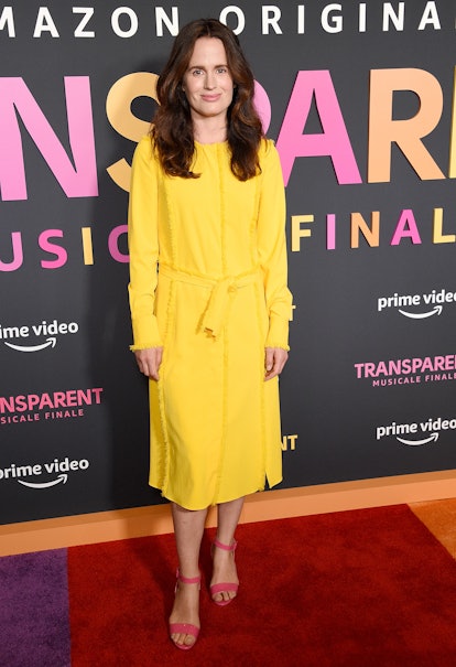 Elizabeth Reaser stands on the red carpet, smiling. She is wearing a yellow dress and a pair of pink...