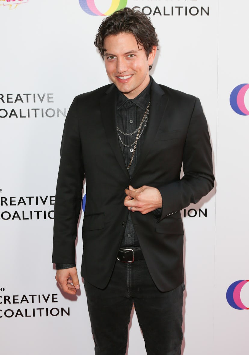 Jackson Rathbone stands on the red carpet, smiling. He wears a black suit, a black pinstripe shirt, ...