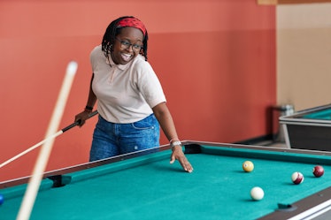 A young Black woman laughs while playing pool in a college collab house.
