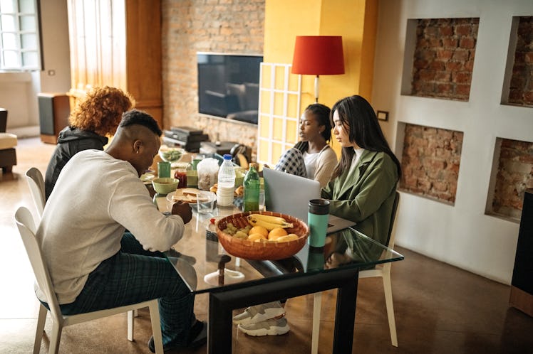 A group of students hangs out at a kitchen table in a college collab house, with food and their lapt...
