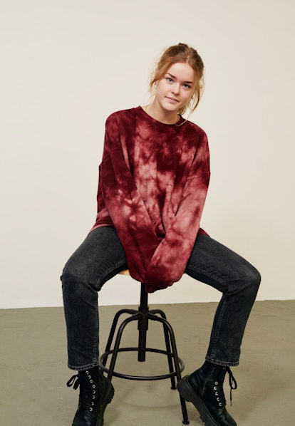 A young woman poses on a stool while wearing a tie-dye crewneck sweatshirt, black jeans, and combat ...
